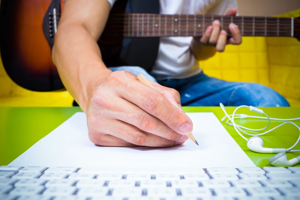 Person composing music and getting paid to write song lyrics while playing the guitar. Close up of a hand writing on paper with a computer keyboard in the foreground. In the background we can see a guitar on their lap. The person's face is not visible.