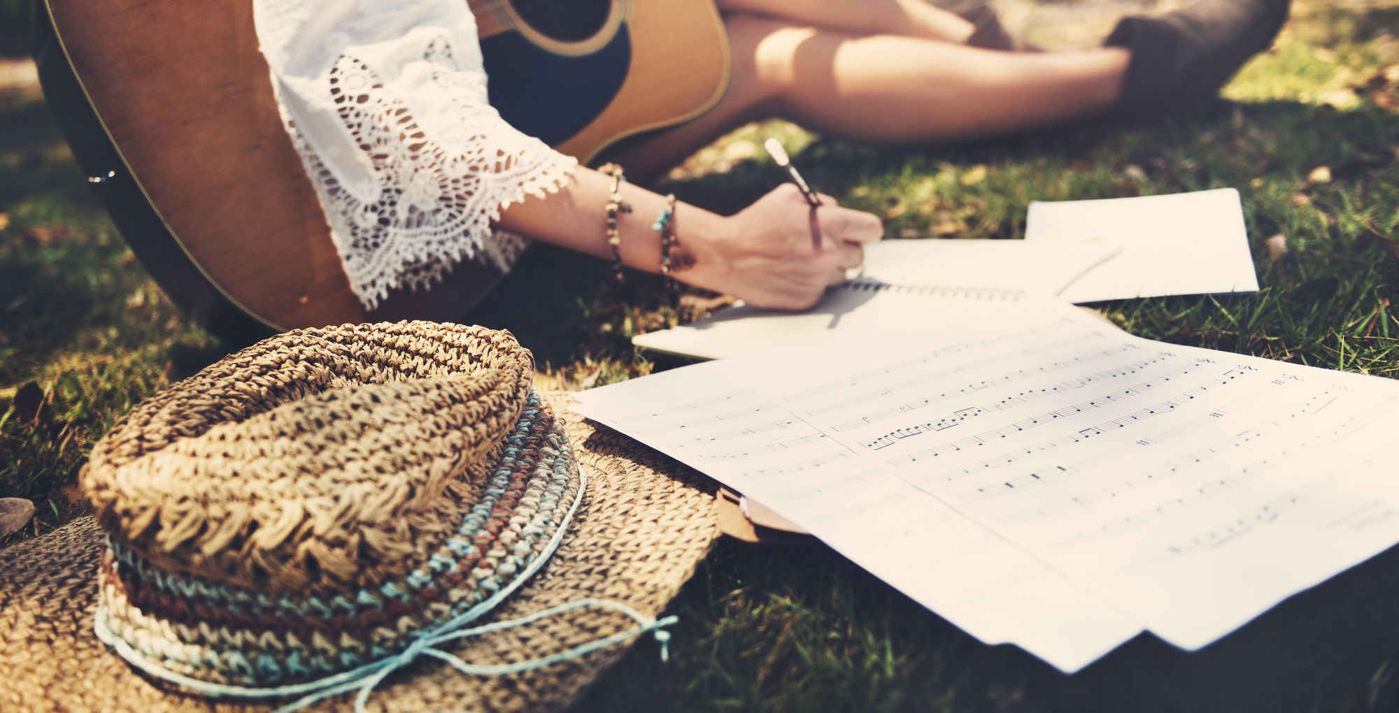 A young female songwriter lying on the grass holding a guitar and writing sheet music