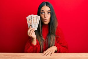 Beautiful dark haired woman holding several £10 notes fanned out, looking amazed with open mouth for surprise, red background