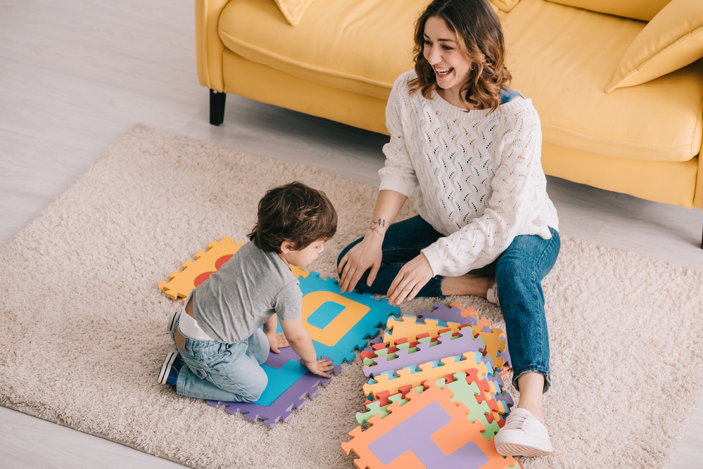 A smiling woman and small child sit on the floor playing with foam tiles with letters on. Babysitting or childminding is a great way to earn extra cash in the UK.