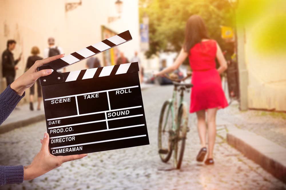 how to make extra money uk as a movie extra. A film set on a city street. Hands holding a clapperboard in the foreground, a woman in a red dress pushes a bicycle behind.