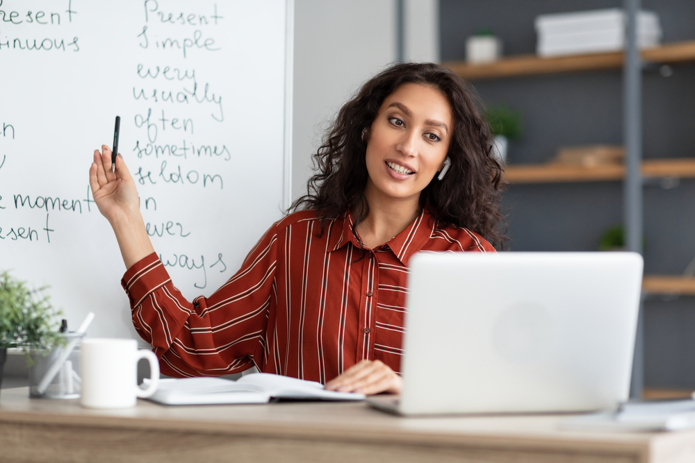 a young woman sitting at a laptop, pointing to a whiteboard behind her on which English vocabulary items are written. Teaching English online is a good way to make extra money in the UK.