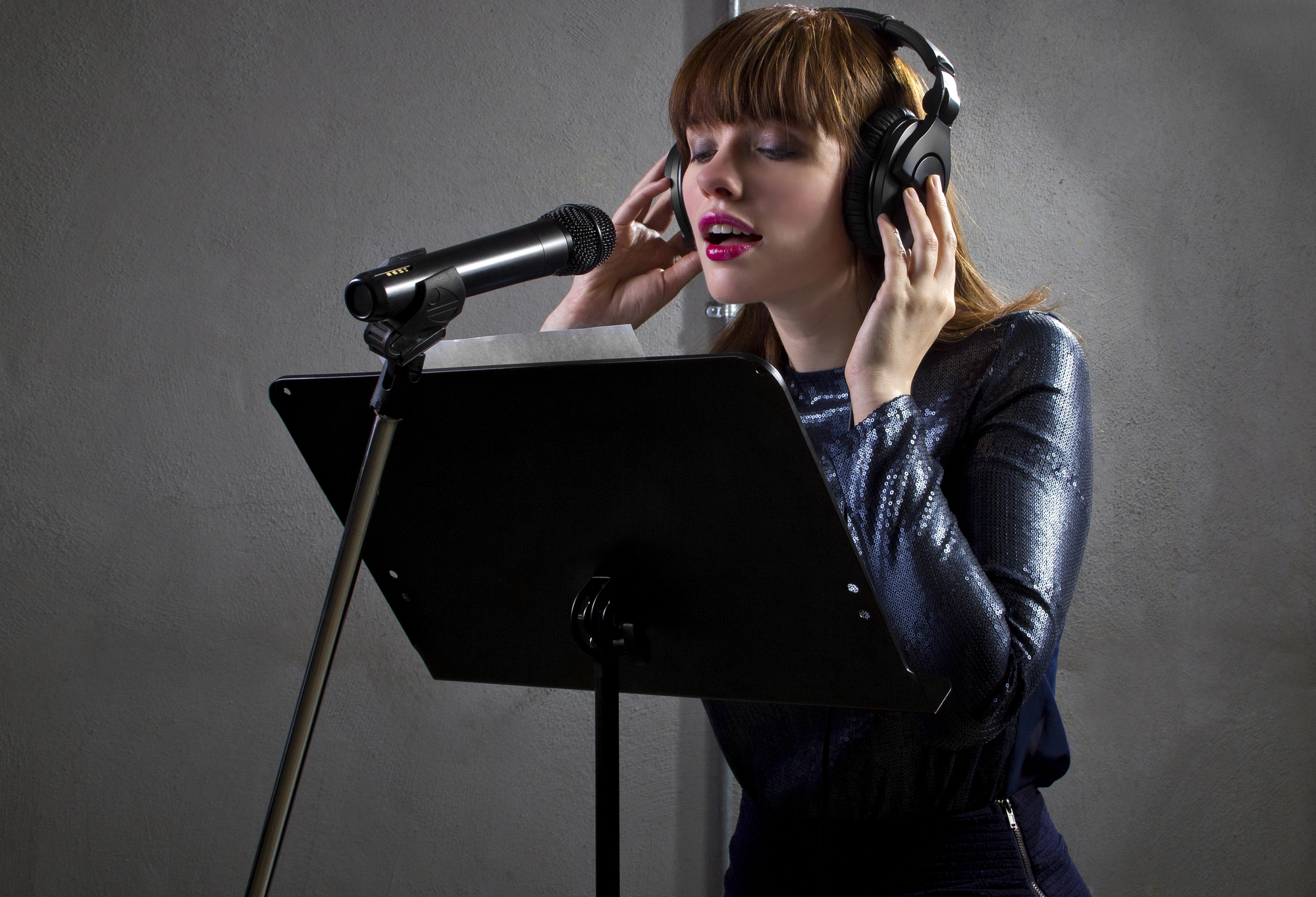 Female professional singer/songwriter in headphones singing into a microphone with a music stand in front, against a dark background