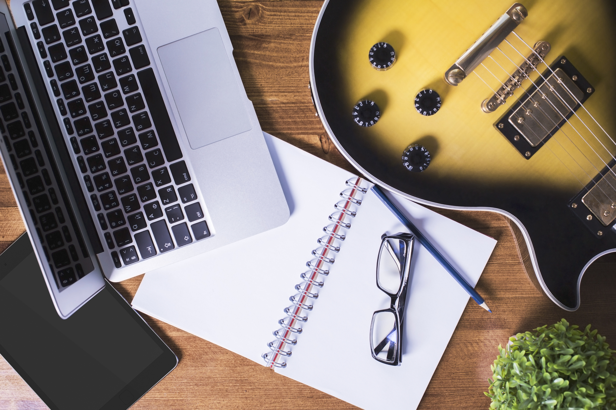Flatlay of a laptop, guitar, notebook, glasses, and a pencil on a wooden desk suggesting a creative workspace where one can write song lyrics or engage in music composition.