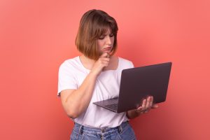 young woman in white t shirt and blue jeans against light red background, holding laptop and looking worriedly at the screen, wondering is matched betting safe?