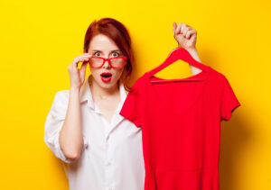 Young woman with red glasses holding a red T-shirt on a hanger against a bright yellow background, learning how to make money online selling t shirts