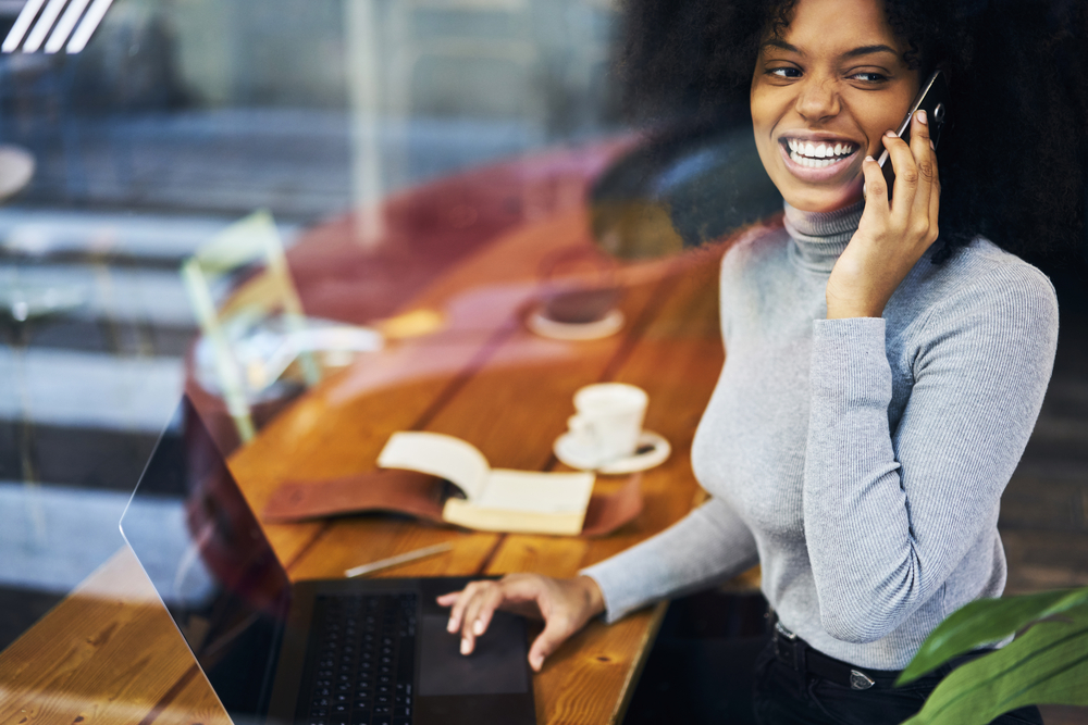 Woman on phone smiling sitting in front of laptop with open notebook.