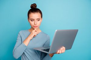 Young woman in blue against blue background, looking puzzled at her laptop that she is holding in her hands, considering no risk matched betting