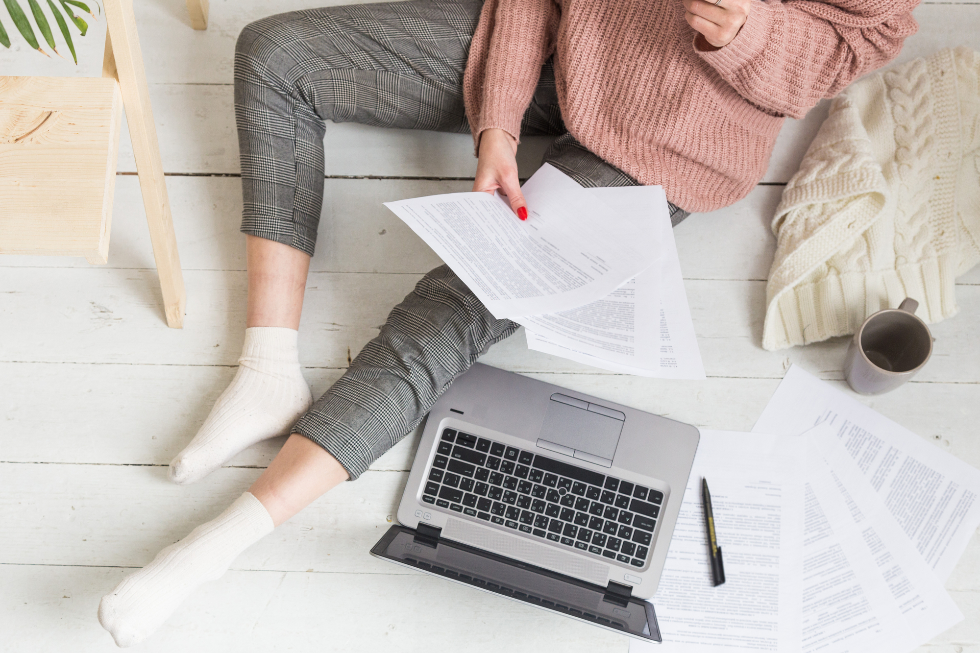 Female student in pink top and grey leggings sits on the floor with her laptop and several sheets of paper, working on her freelance writing side hustle student job