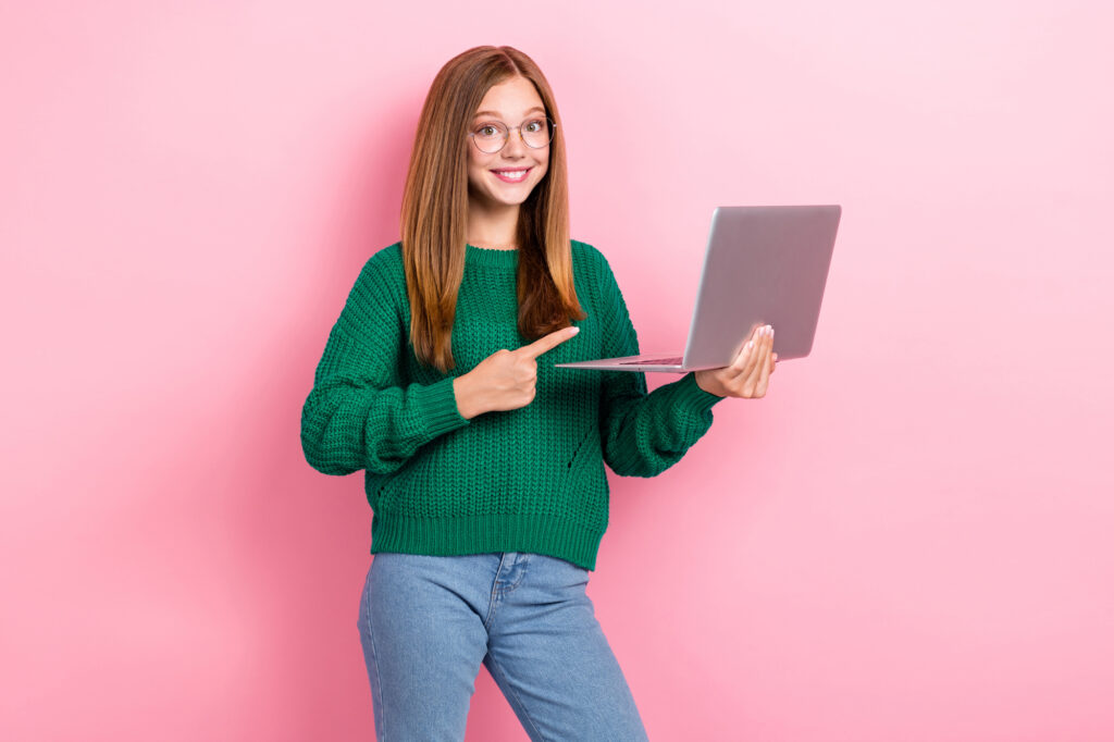 Teenage girl in glasses and green sweater pointing at laptop screen, smiling, standing against a pink background, searching for online jobs for teens.