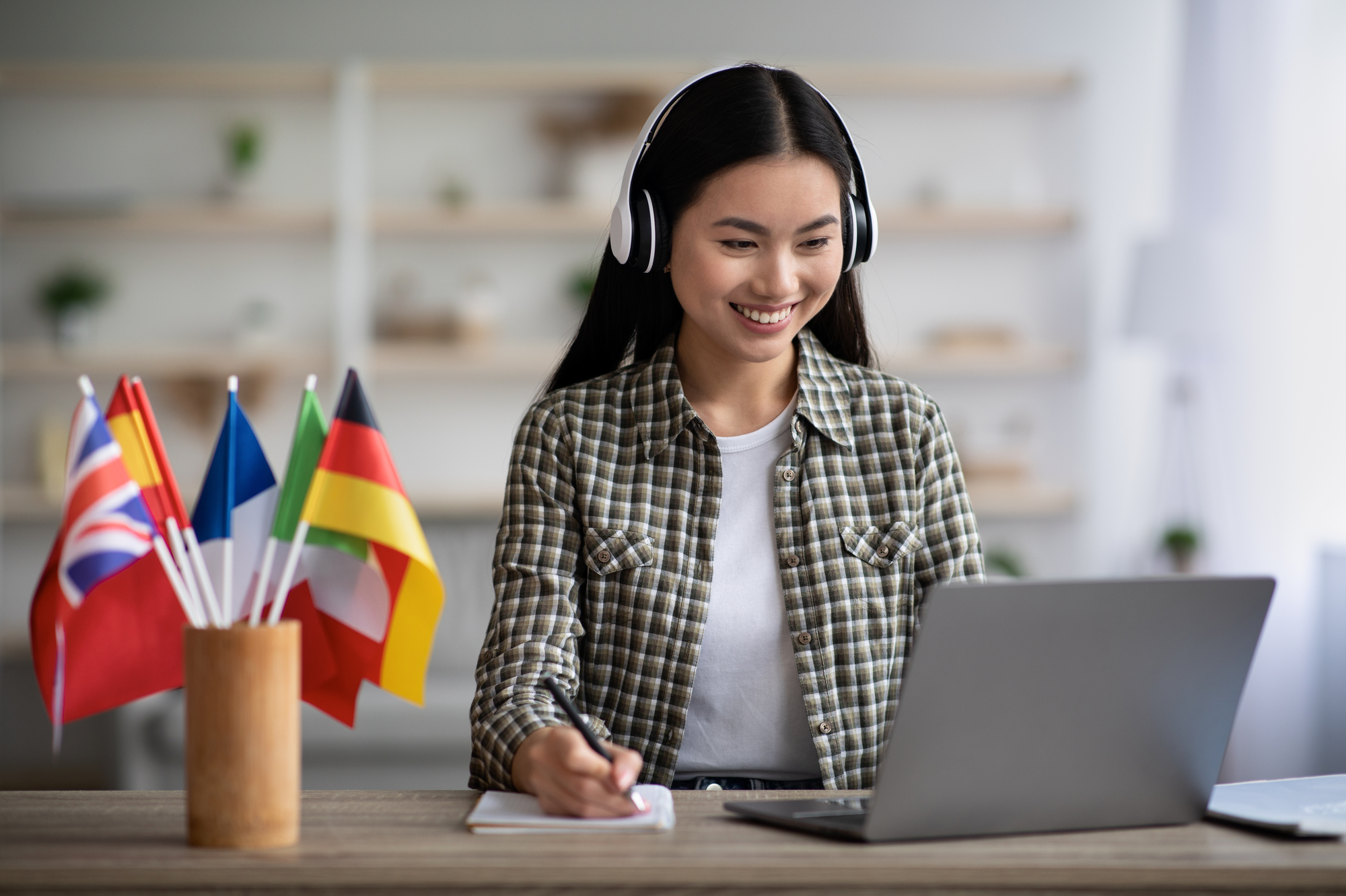 A woman with headphones is sitting at a desk with international flags in front of her, teaching English as her online student job.