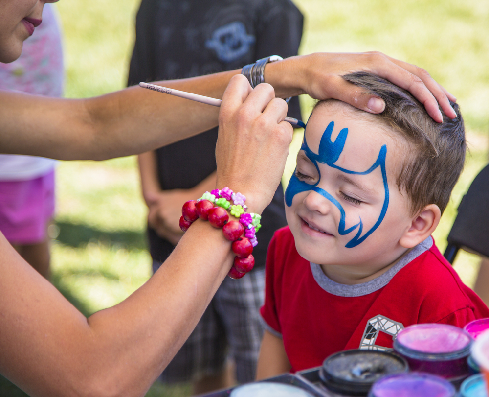 A woman in the UK engaging in a face painting side hustle by painting a child's face with blue paint in the shape of a batman symbol.
