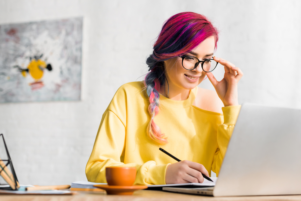 A woman in glasses with pink hair is working on her laptop at a desk, managing her ebook writing side hustle in the UK.
