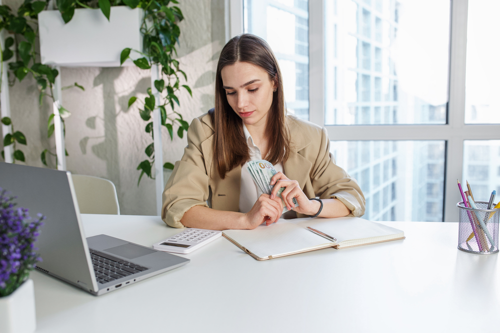 A woman is sitting at a desk researching funding options for her side hustle using a laptop.