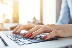 Close-up of a woman's hands with red nail polish typing on a laptop keyboard, illuminated by natural light, researching how to make money online.