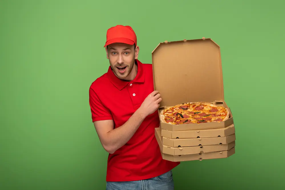 a happy pizza delivery man in red with a stack of pizzas, showing an open pizza box