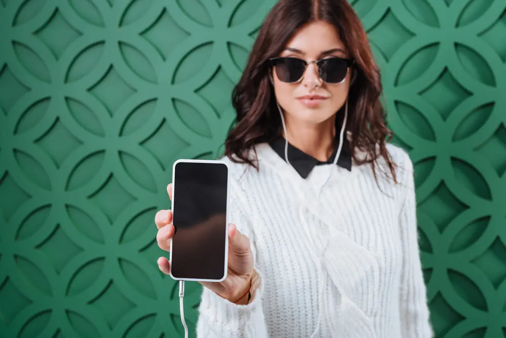 woman in sunglasses holding out mobile phone; green background