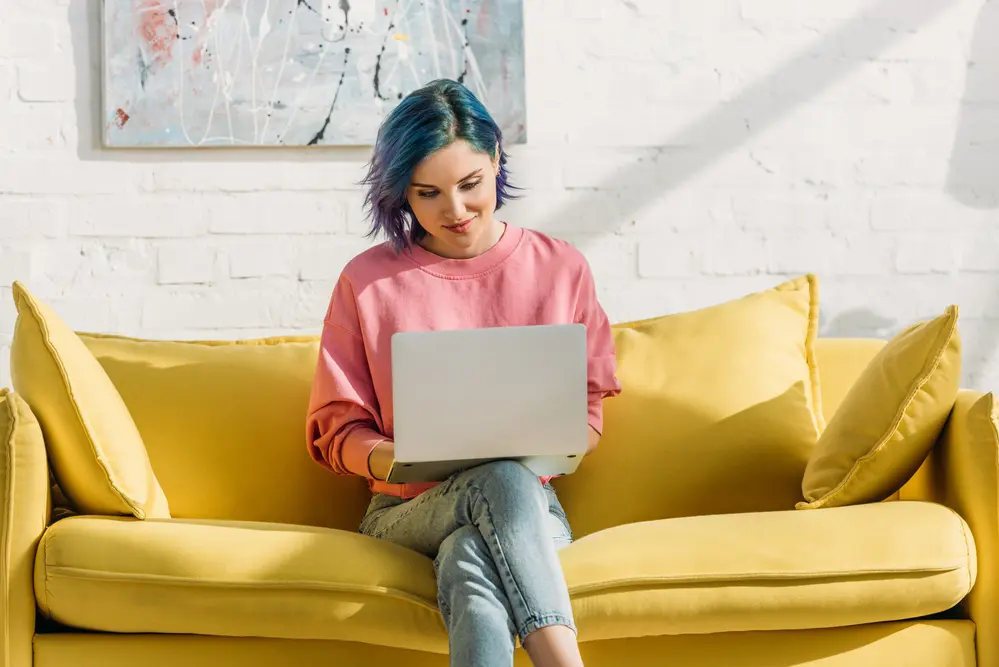 a woman with blue hair on a yellow sofa working on gpt sites on her laptop