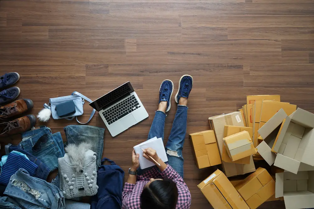 A woman sits with a laptop and notepad, surrounded by piles of shipping boxes, working on her Amazon business.