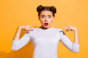 a woman on yellow background looking shocked and pointing at herself