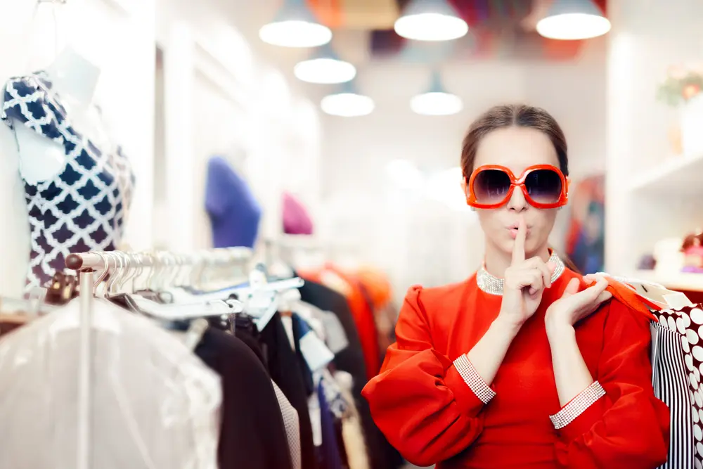 a mystery shopper wearing big sunglasses and holding a finger to her lips, in a clothes shop