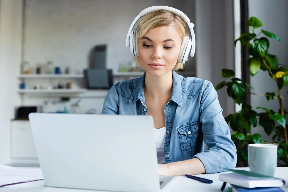 a woman in blue denim shirt sitting in front of her laptop, wearing headphones