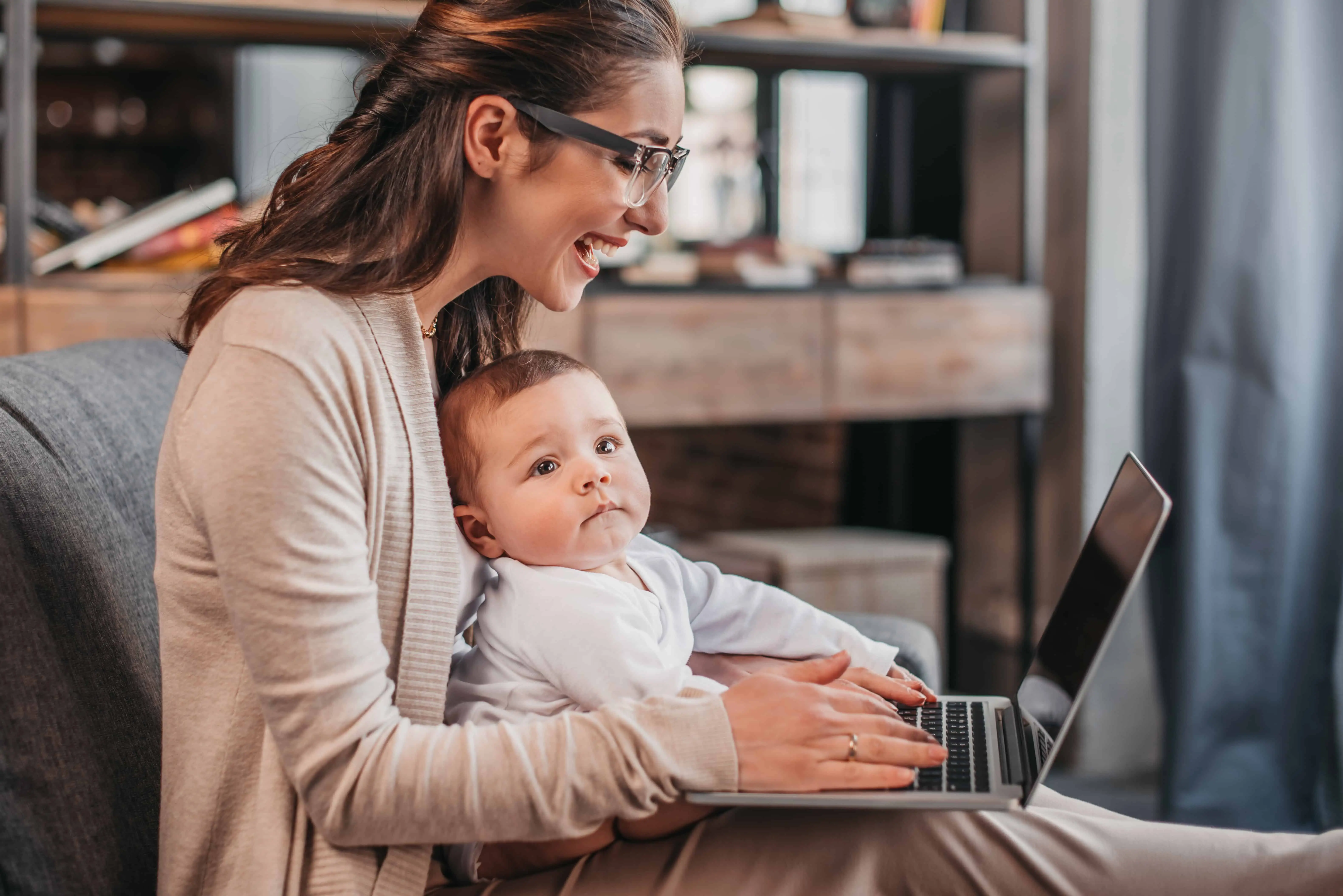 work from home jobs for mums - a woman working from home at a laptop holding a baby